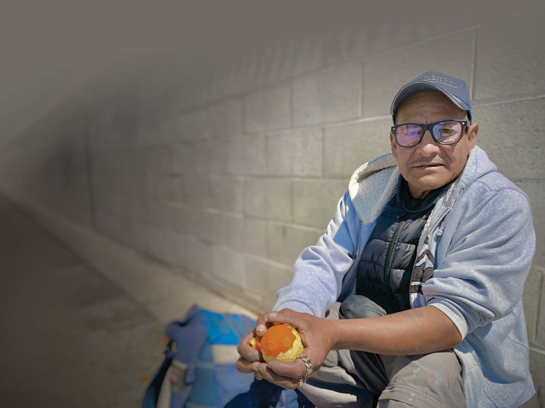 A person wearing glasses and a cap sits against a brick wall, holding an apple, contemplating how they might donate to those in need for Christmas 2024.