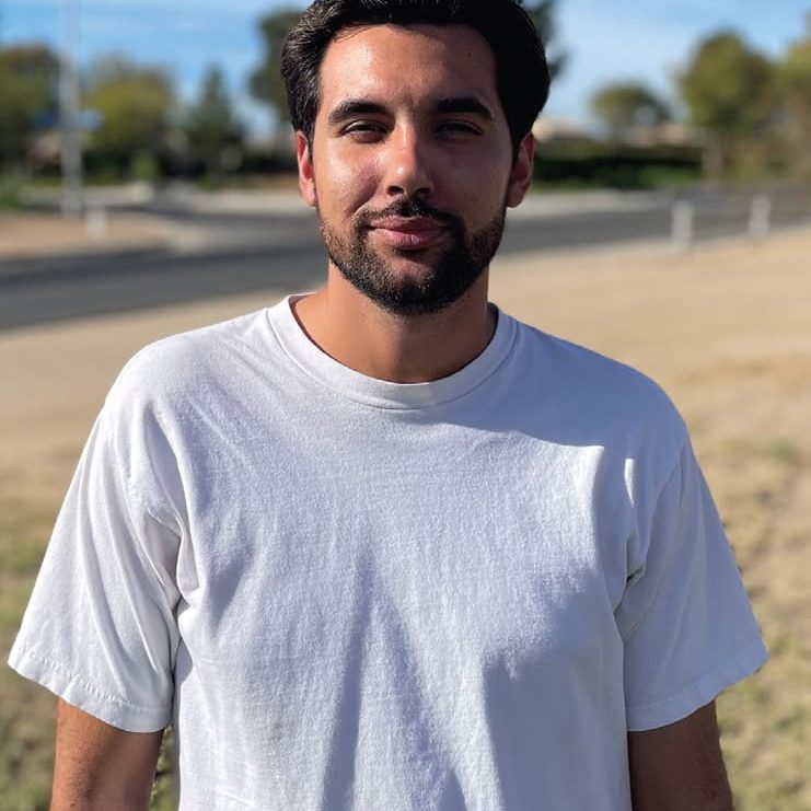 A person with dark hair and a beard stands outdoors in Victorville, wearing a white T-shirt. The background includes a road and trees under a clear sky.